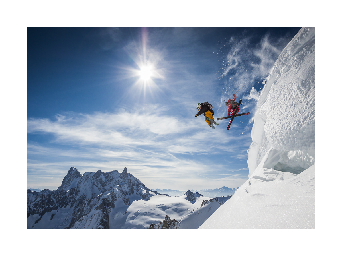 Zwei Skifahrer springen von einem Schneehügel vor einem klaren blauen Himmel und schneebedeckten Bergen.