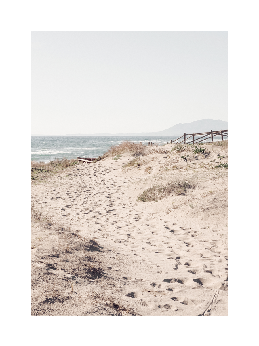 Fotografie eines Sandweges mit Fußspuren, der zu einem Strand führt, mit Meer und Bergen im Hintergrund.