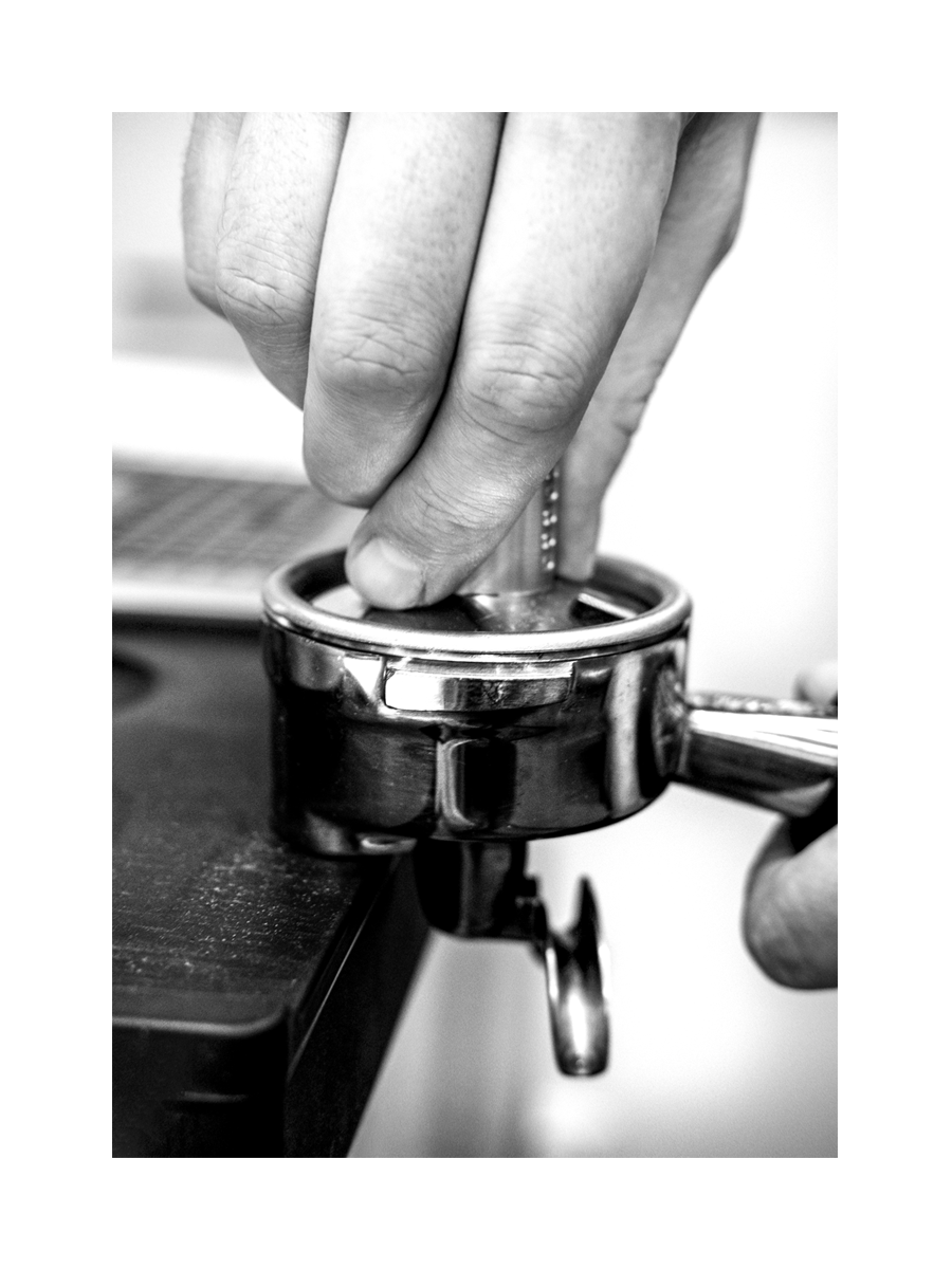 Schwarz-Weiß-Fotografie einer Hand, die Kaffeepulver in einen Siebträger drückt.