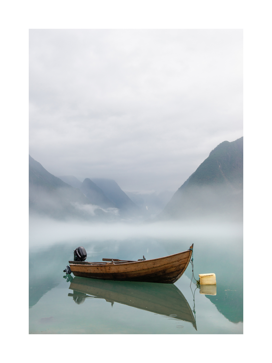 Fotografie eines Holzboots auf einem nebligen See mit Bergen im Hintergrund.