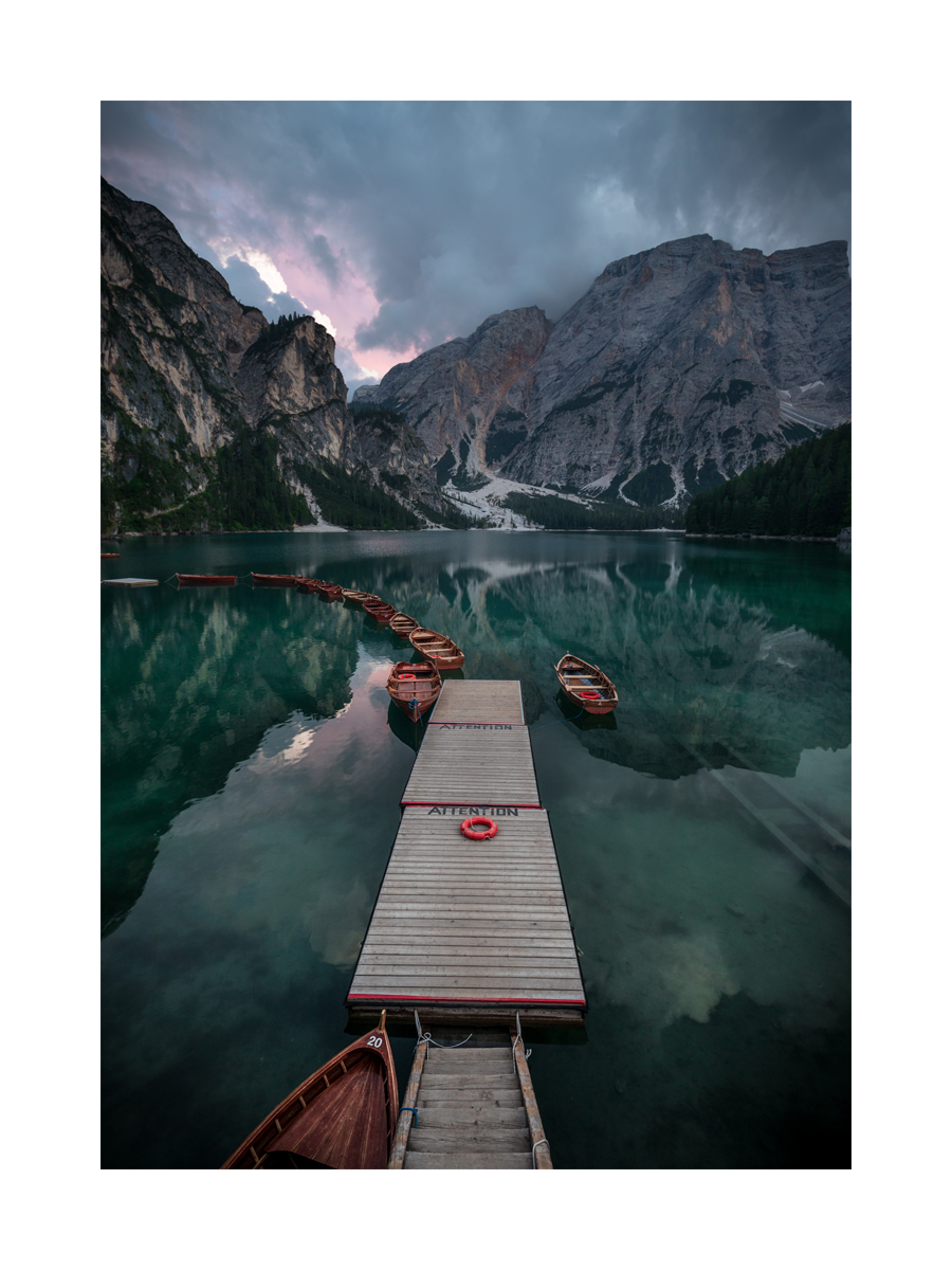 Fotografie eines Stegs mit Booten auf einem See und Bergen im Hintergrund.