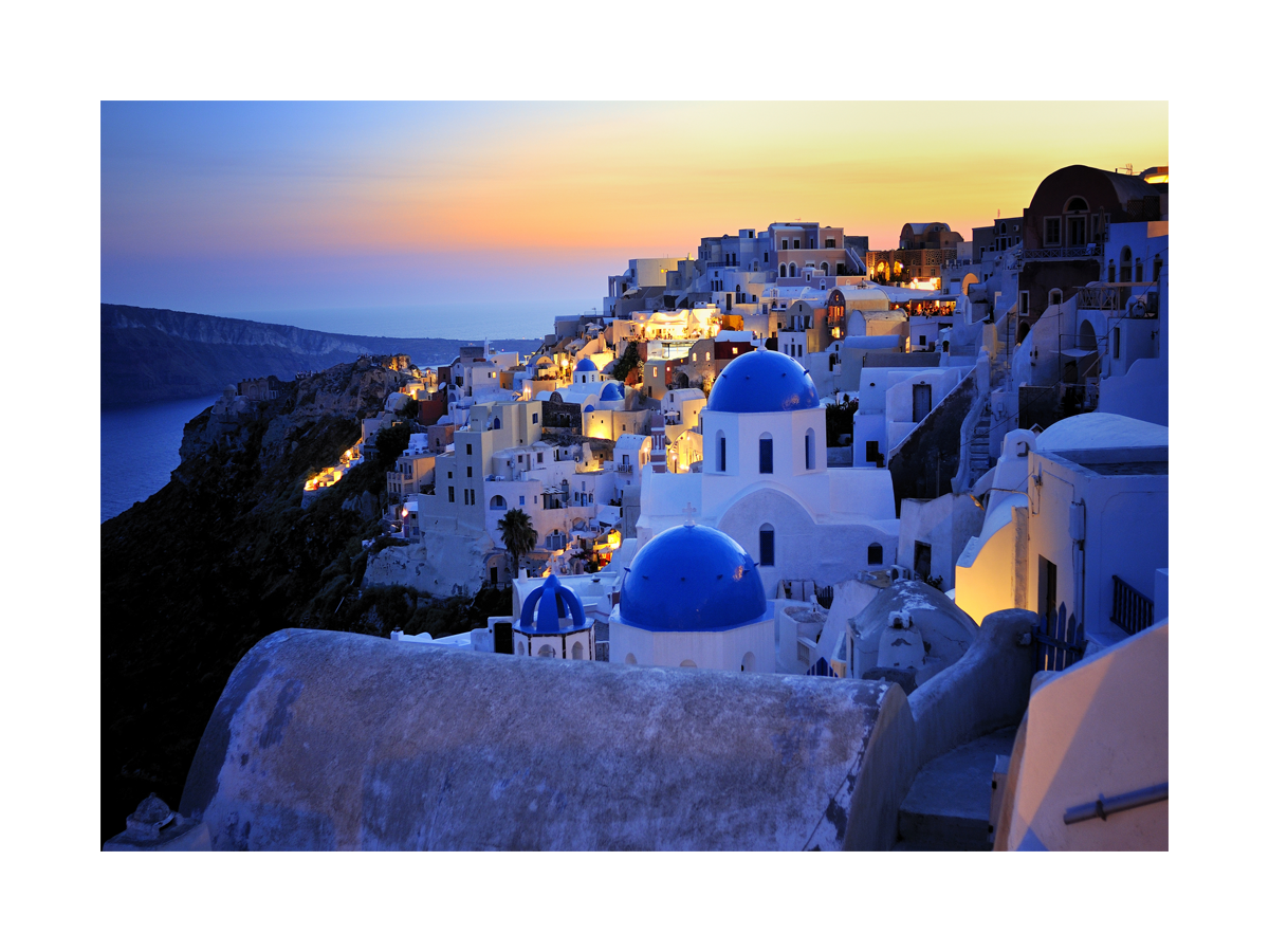 Fotografie der Stadt Oia auf Santorini bei Sonnenuntergang mit dem Meer im Hintergrund.