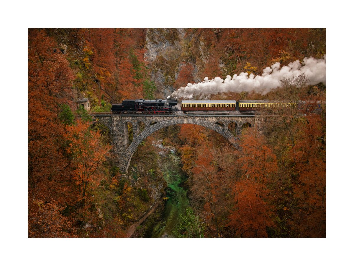 Ein Zug mit gelben Waggons überquert eine steinerne Brücke inmitten von Herbstbäumen.