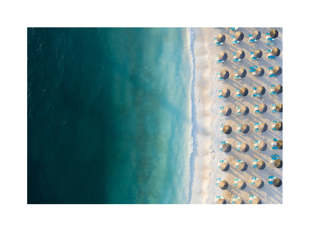 Luftaufnahme eines Strandes mit Sonnenschirmen und türkisfarbenem Wasser.