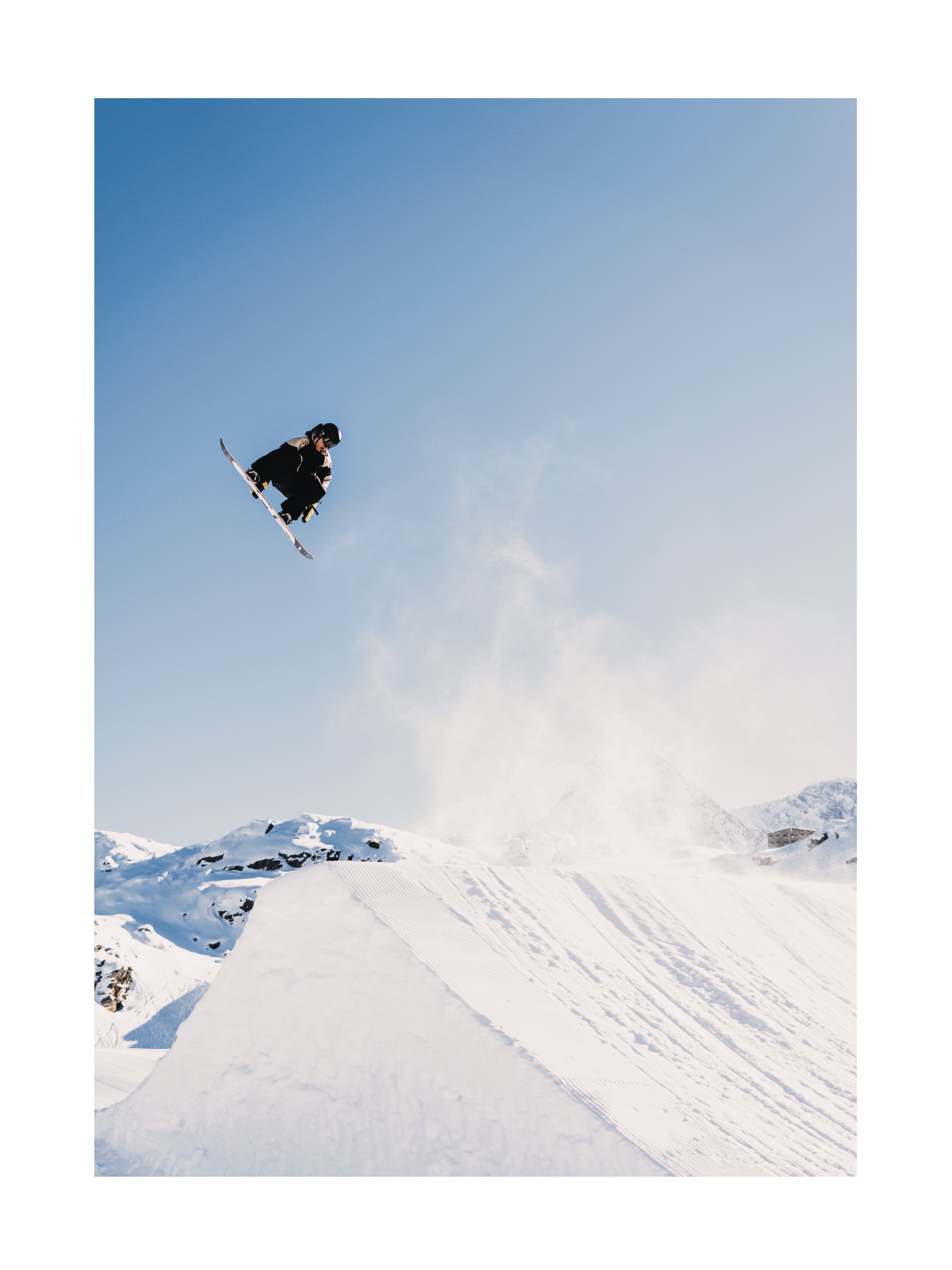 Ein Snowboarder springt in der Luft vor einem blauen Himmel und schneebedeckten Bergen.