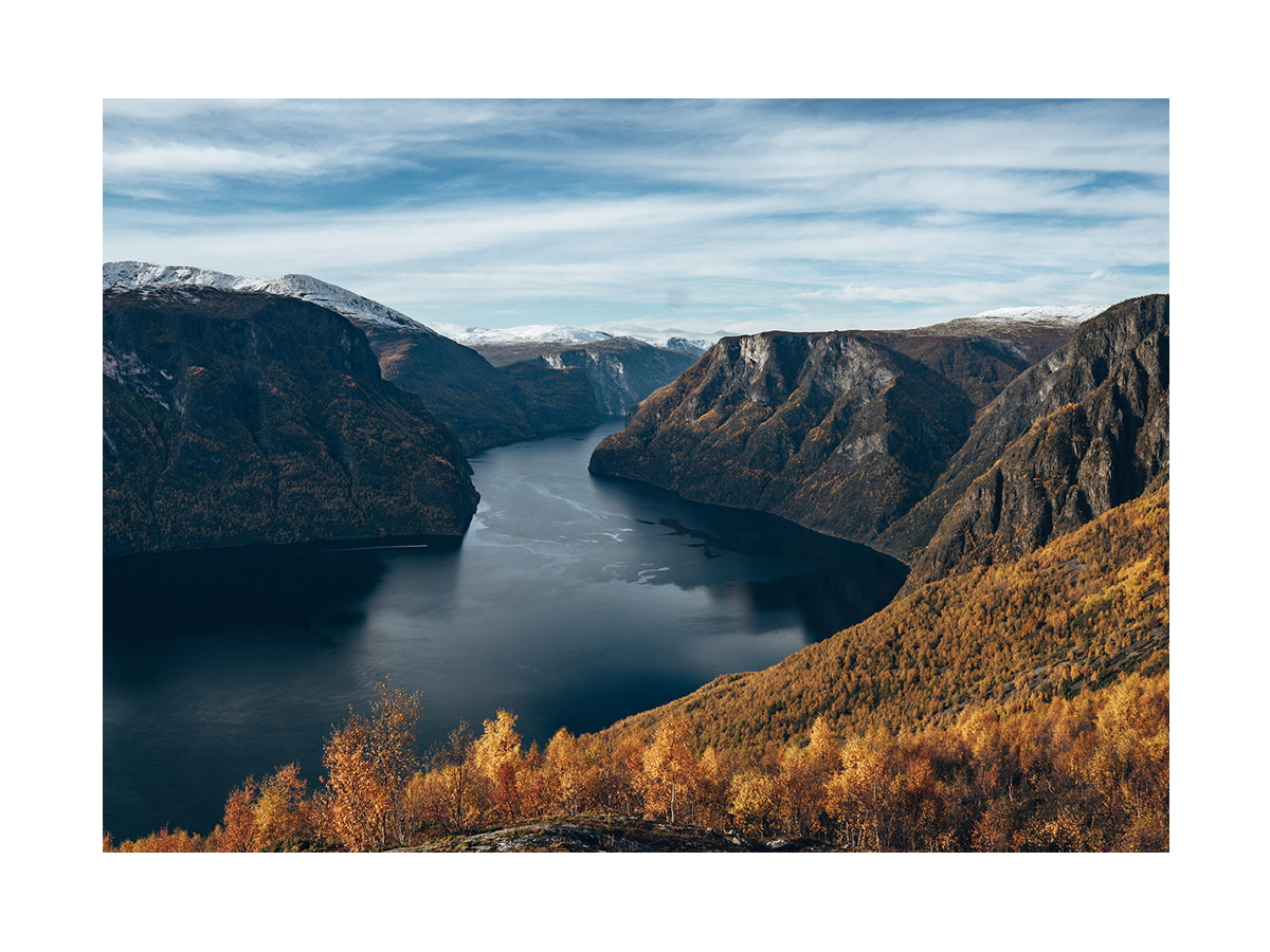 Fotografie eines Fjords mit schneebedeckten Bergen und herbstlichen Bäumen.