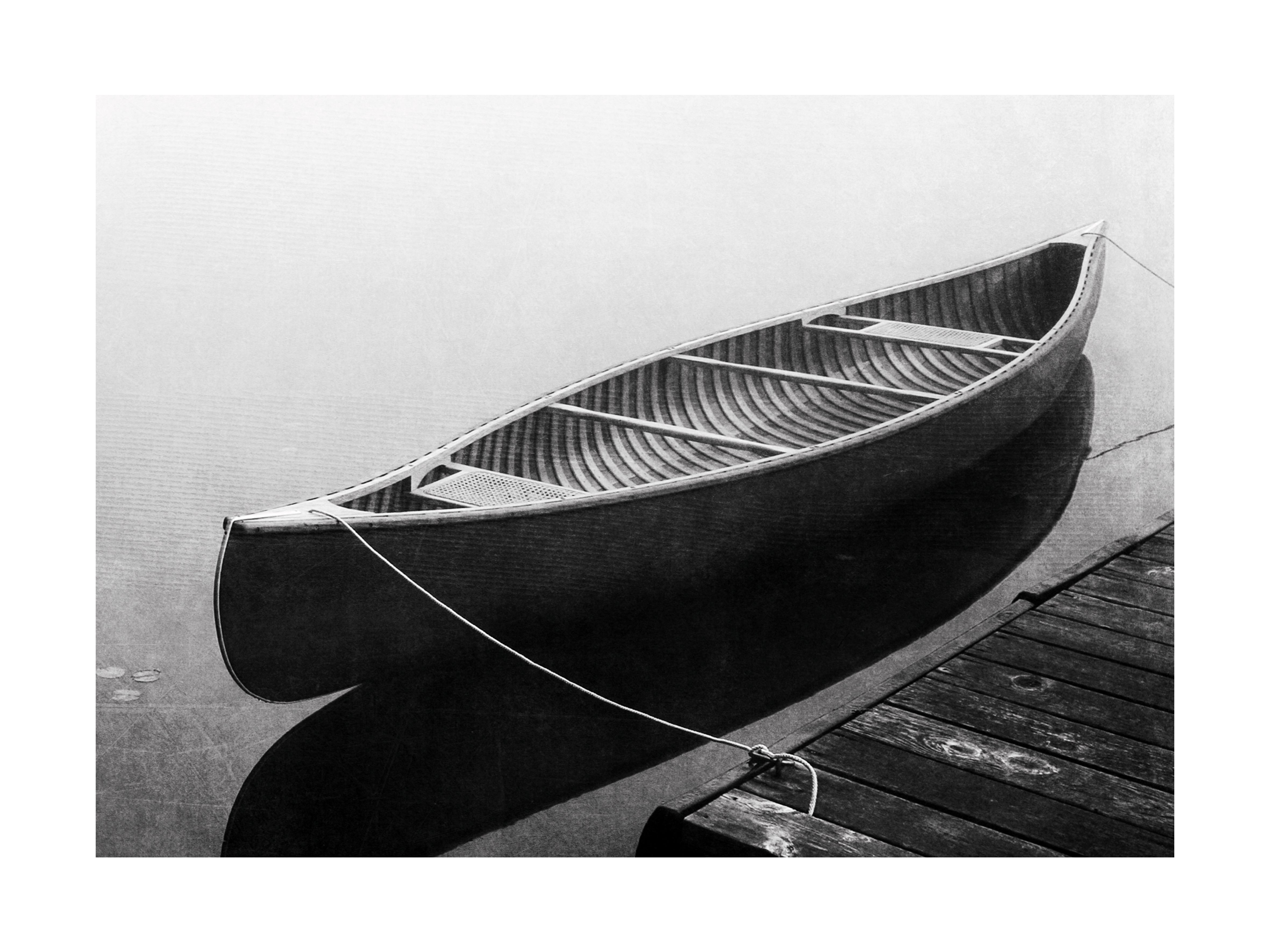 Schwarz-weißes Foto eines Kanus an einem Steg auf ruhigem Wasser.