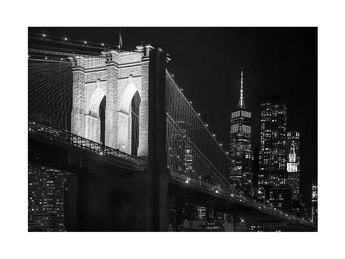Schwarz-Weiß-Fotografie der Brooklyn Bridge bei Nacht mit einem Wolkenkratzer im Hintergrund.