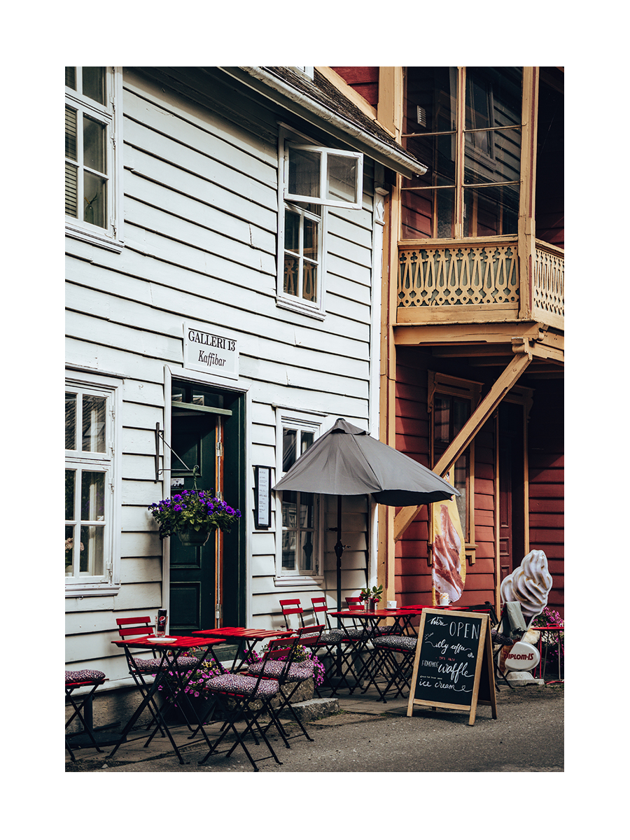 Fotografie eines norwegischen Cafés mit roten Stühlen und grauem Sonnenschirm vor einem weißen Gebäude.
