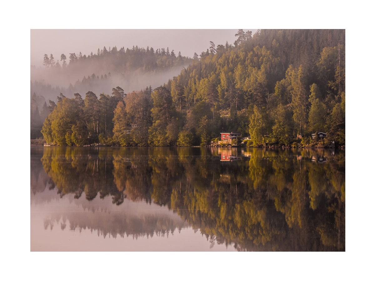 Ein rotes Haus spiegelt sich in einem ruhigen See, umgeben von grünen Bäumen und Nebel.