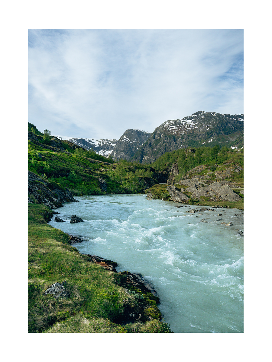 Ein Fluss in Norwegen, umgeben von grüner Landschaft und schneebedeckten Bergen unter blauem Himmel.