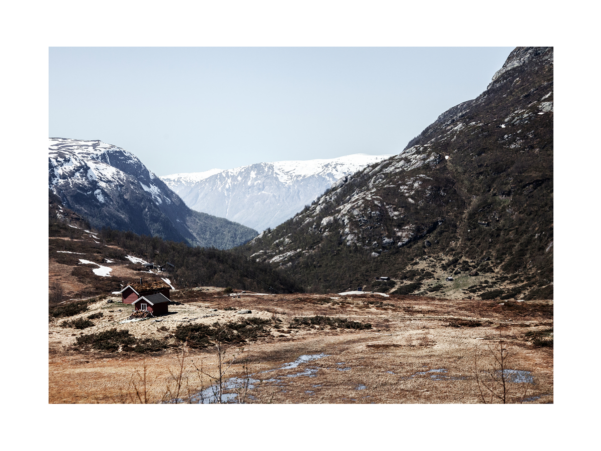 Fotografie einer rotbraunen Hütte in einem Tal mit schneebedeckten Bergen im Hintergrund.