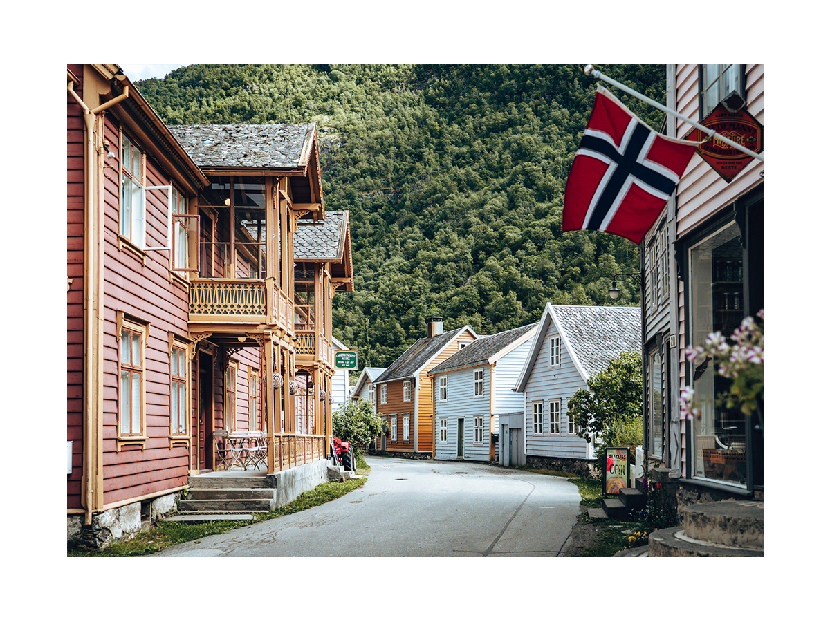Fotografie eines norwegischen Dorfes mit bunten Holzhäusern und einer norwegischen Flagge, umgeben von grünen Bergen.