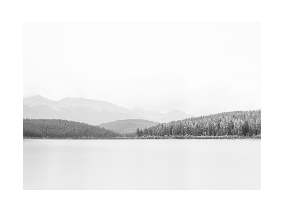 Schwarz-Weiß-Fotografie eines Sees mit Wald und Bergen im Hintergrund.