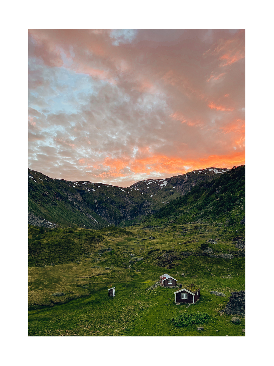 Fotografie einer Hütte in einer Berglandschaft bei Sonnenuntergang mit orangefarbenem Himmel.