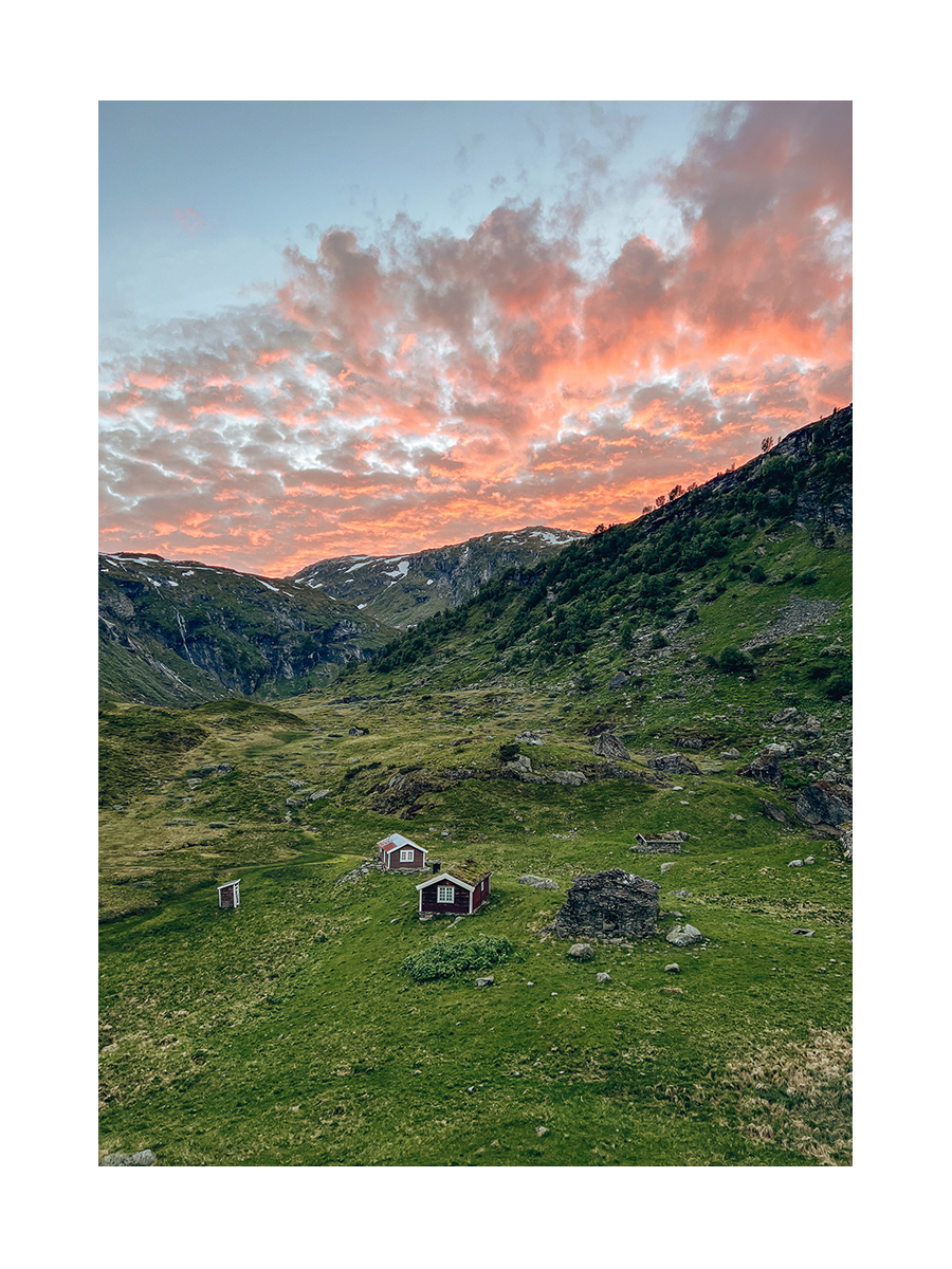 Landschaftsfoto mit grünen Hügeln, roten Häusern und einem orangefarbenen Himmel.