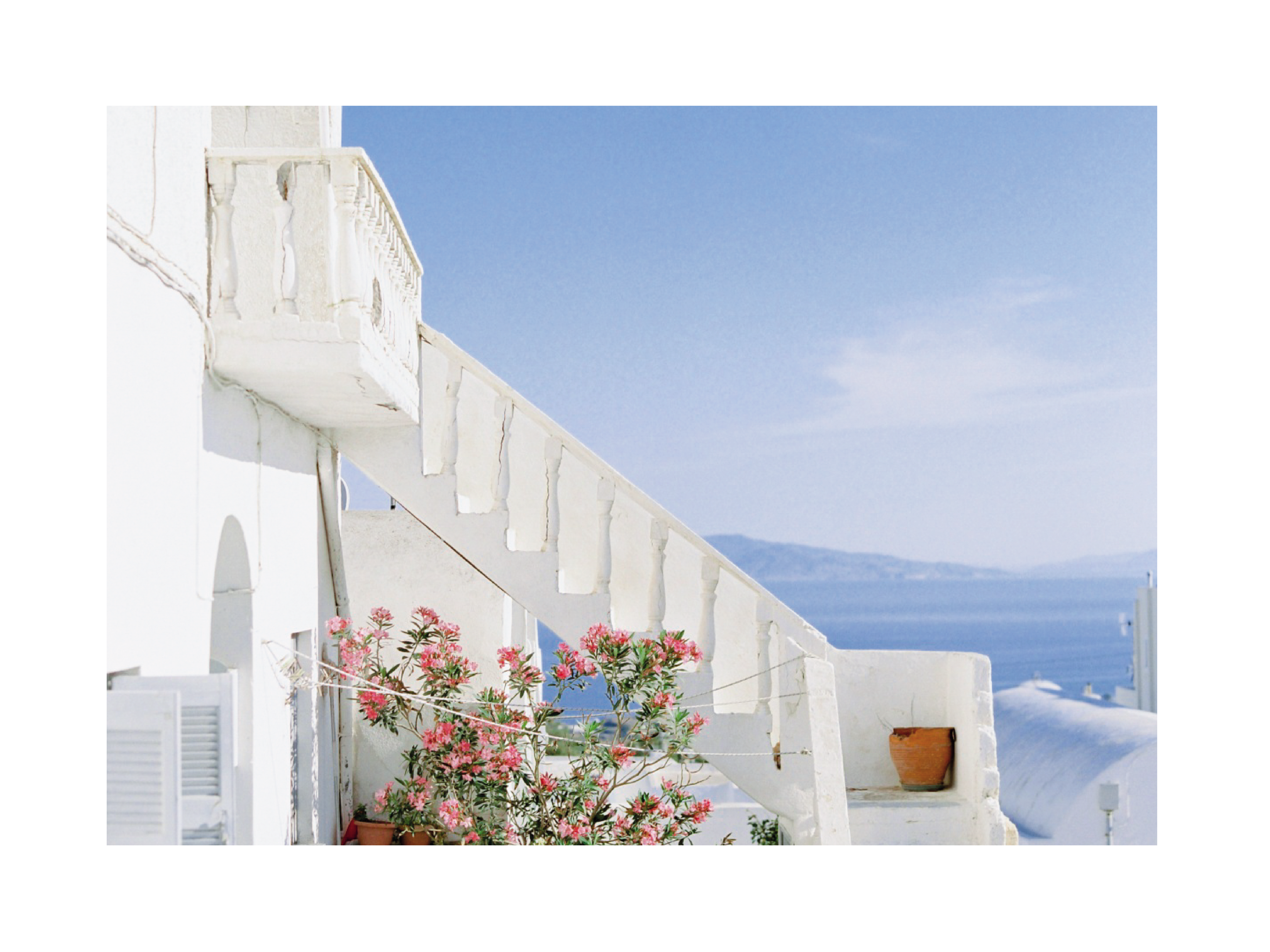 Fotografie einer weißen Treppe mit rosa Blumen und Meer im Hintergrund.