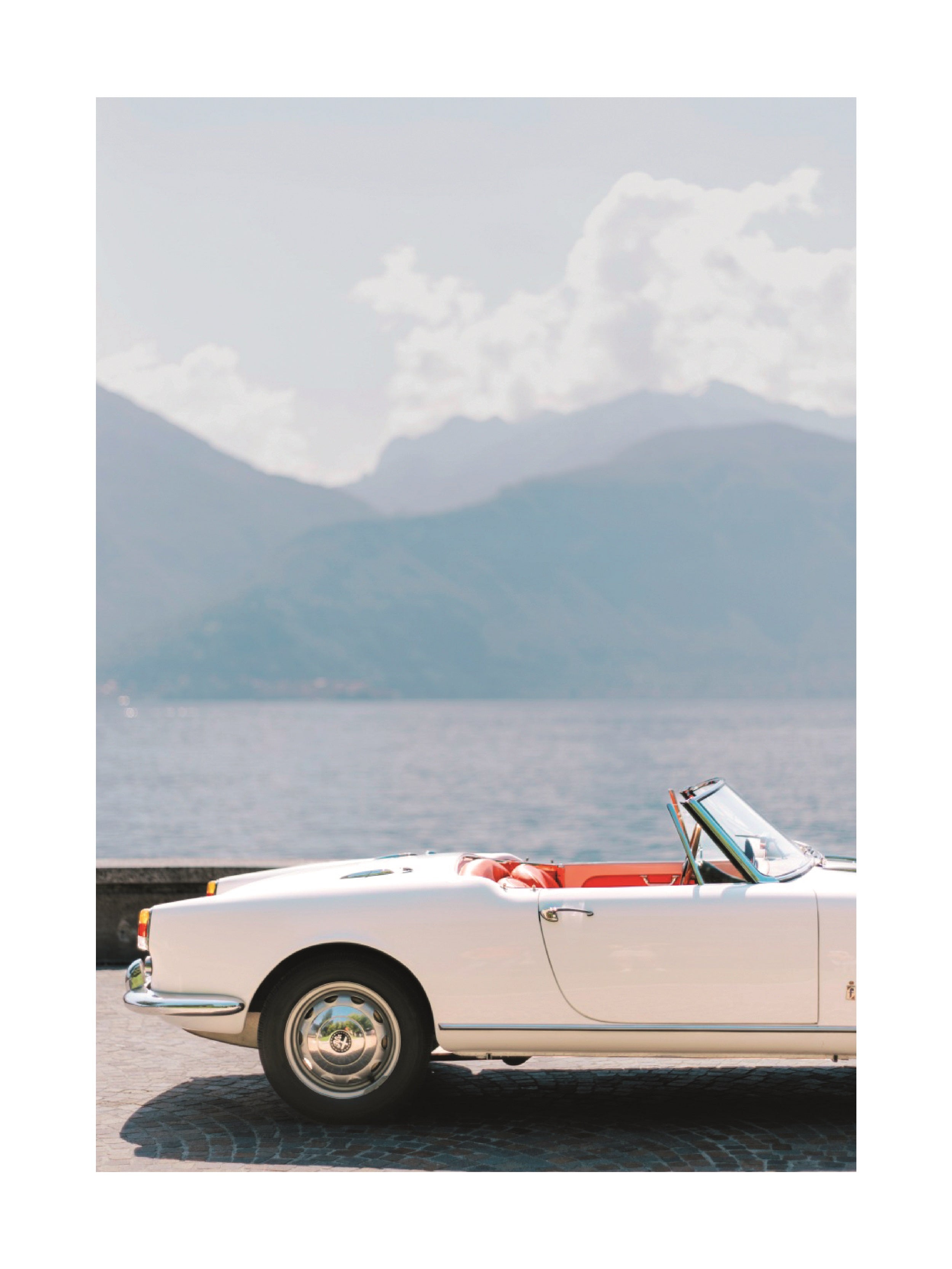 Fotografie eines weißen Cabriolets mit roter Innenausstattung vor einem See und Bergen im Hintergrund.