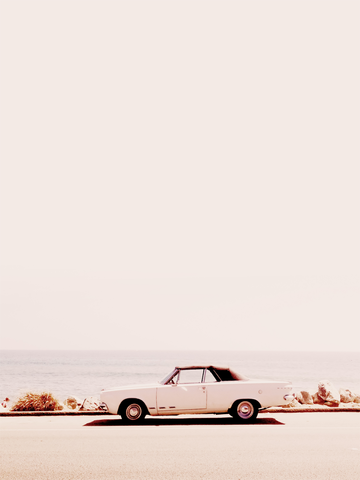 Fotografie eines weißen Cabriolets an einer Küstenstraße mit beigem Himmel.