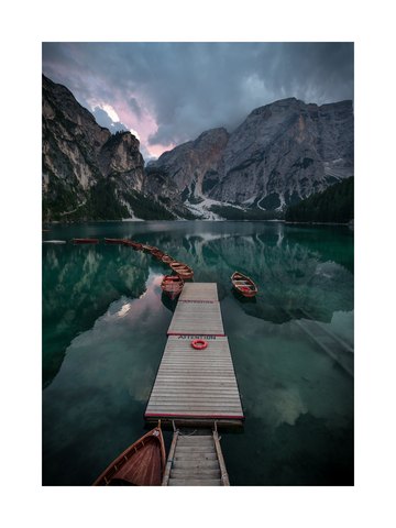 Fotografie eines Stegs mit Booten auf einem See und Bergen im Hintergrund.