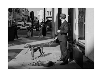 Schwarz-Weiß-Fotografie eines Mannes im Anzug mit zwei Hunden an der Leine auf der Straße.