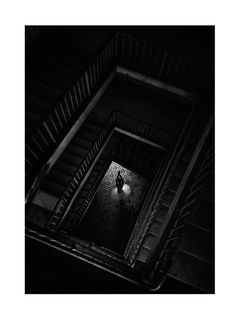 Schwarz-Weiß-Fotografie einer Ballerina in einem dunklen Treppenhaus, von oben beleuchtet.