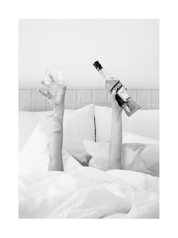 Schwarz-weiß-Fotografie von jemandem, der ein Glas und eine Flasche Aperol im Bett hält.