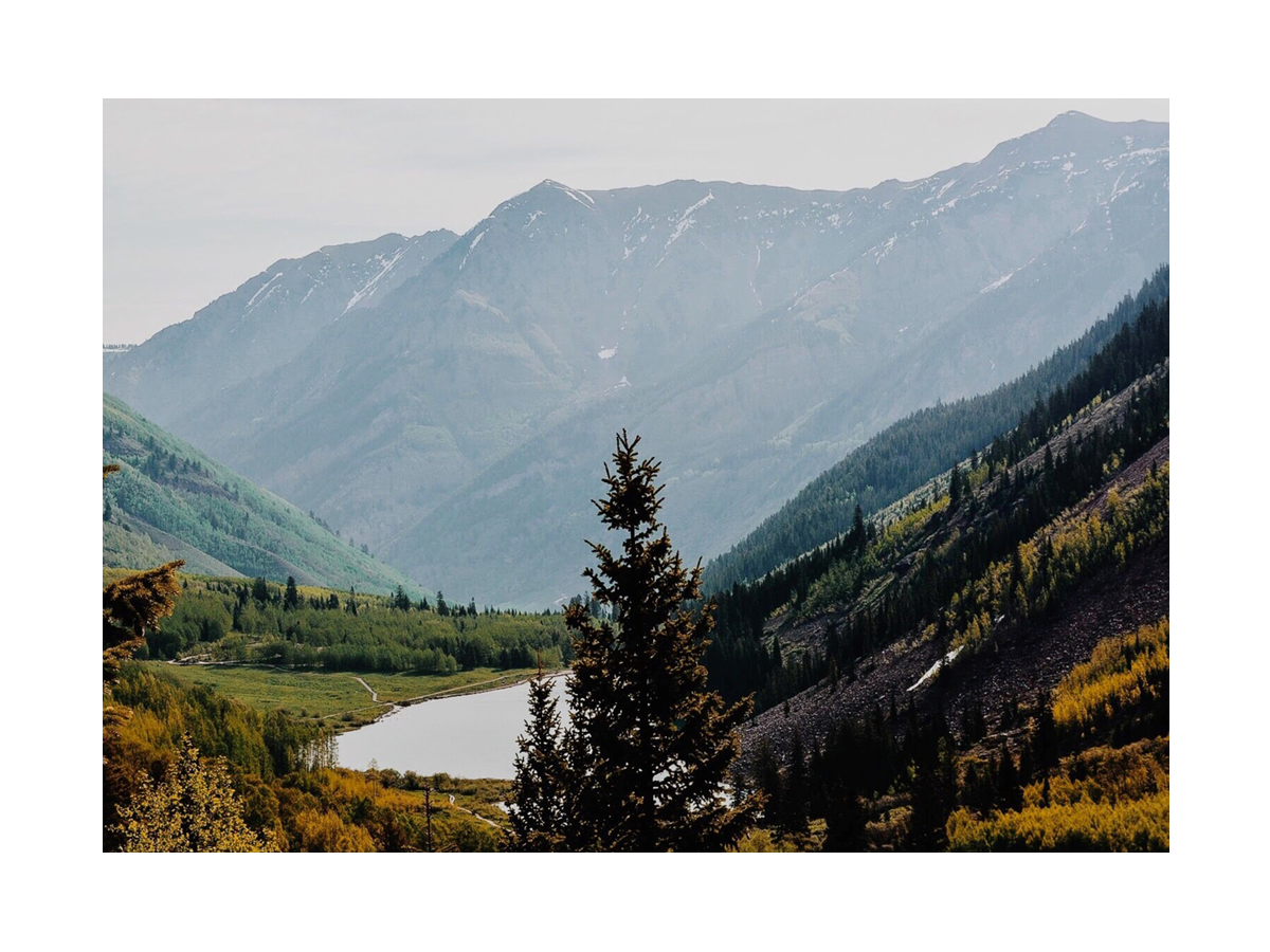 Fotografie einer Berglandschaft mit einem See im Tal und Bäumen im Vordergrund.