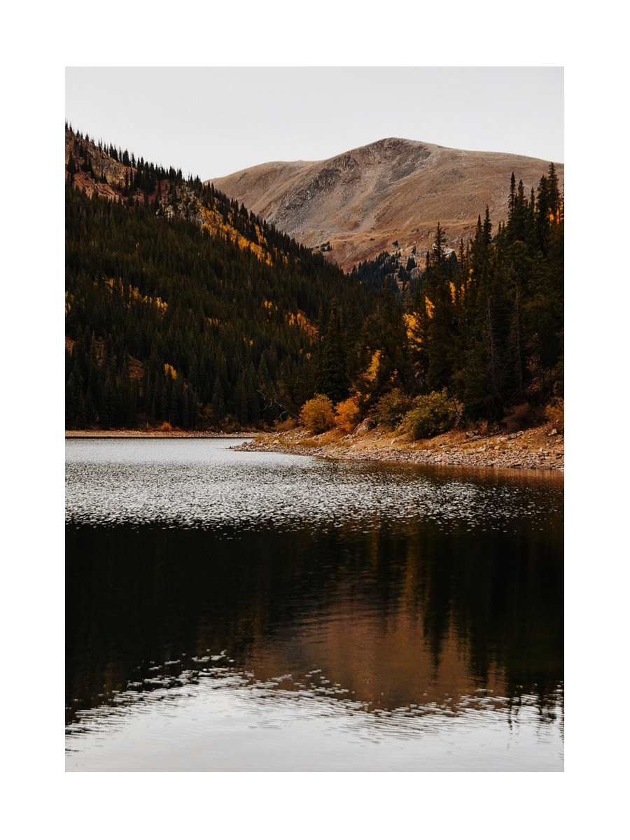 Fotografie eines Sees mit reflektierendem Wasser, umgeben von Nadelwald und Bergen im Herbst.