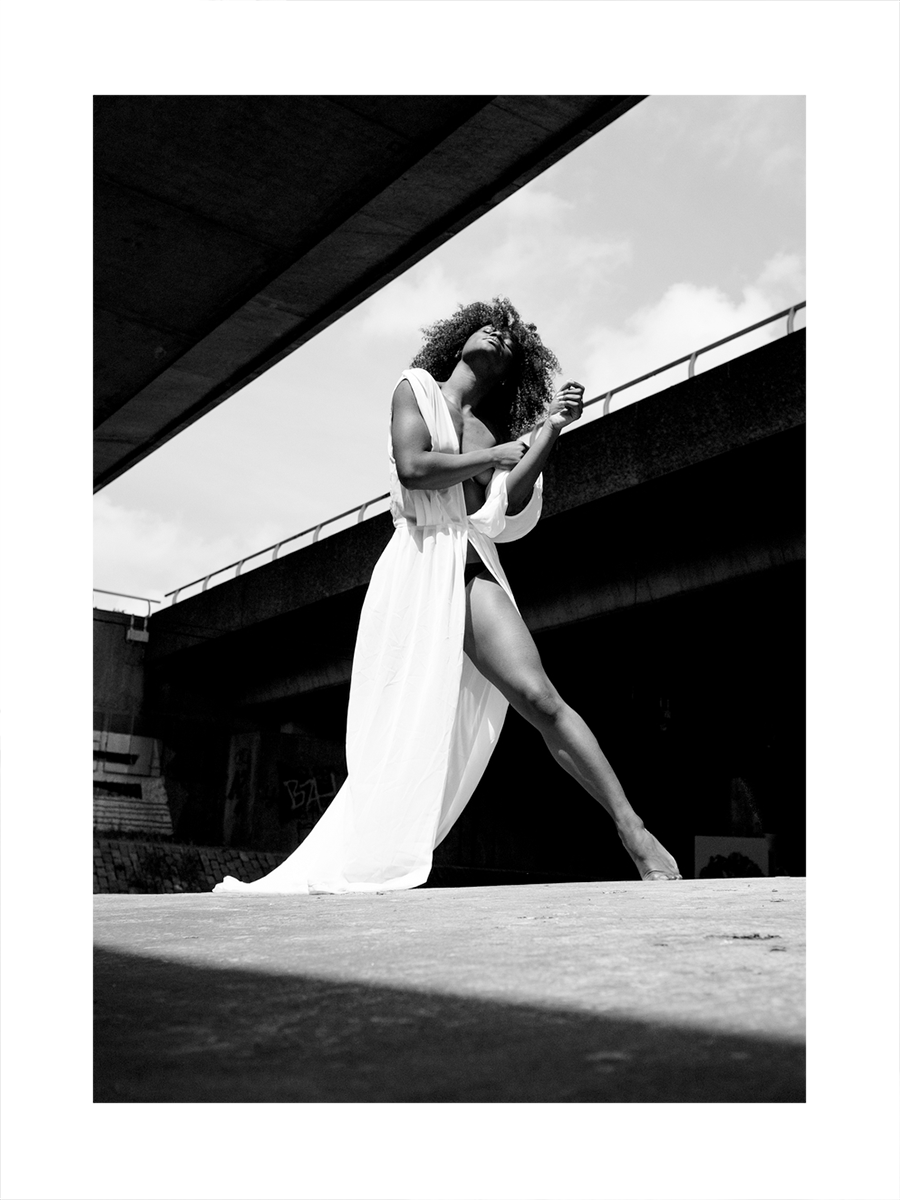 Schwarz-weiß-Fotografie einer Frau in einem weißen Kleid, die unter einer Brücke tanzt.