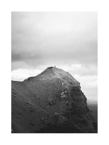 Schwarz-Weiß-Fotografie eines Berggipfels mit einer kleinen Figur und einem bewölkten Himmel.
