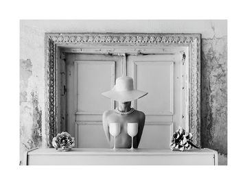 Schwarz-weiß-Fotografie einer Frau mit Hut, die zwei Gläser auf der Brust balanciert, mit Blumen daneben.