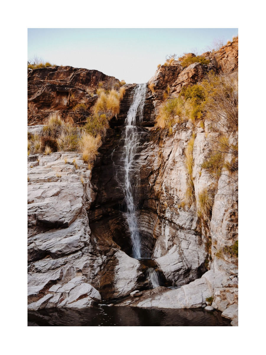 Ein Wasserfall, der in ein ruhiges Wasserbecken fällt, umgeben von Felsen und Pflanzen, mit einem klaren Himmel im Hintergrund.