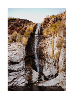 Ein Wasserfall, der in ein ruhiges Wasserbecken fällt, umgeben von Felsen und Pflanzen, mit einem klaren Himmel im Hintergrund.