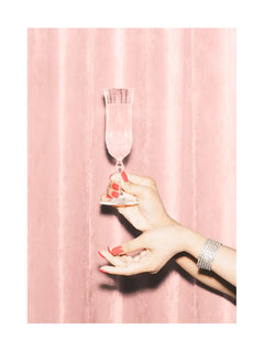 Hand mit roten Nägeln hält ein Champagnerglas vor rosa Hintergrund.