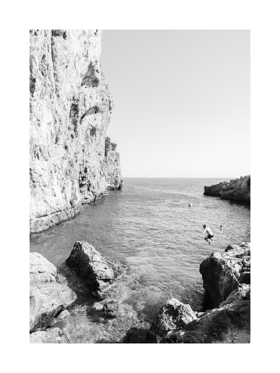 Schwarz-weiß-Fotografie eines Jungen, der von einer Klippe ins Meer springt.