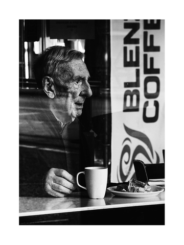 Schwarz-Weiß-Fotografie eines älteren Mannes, der in einem Café sitzt und Kaffee trinkt.