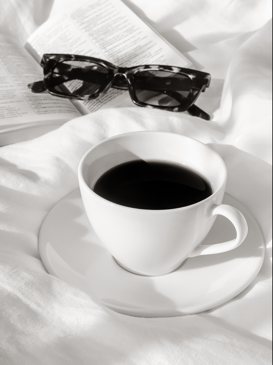 Monochromes Poster mit Kaffeetasse, Buch und Sonnenbrille auf einem Bett.