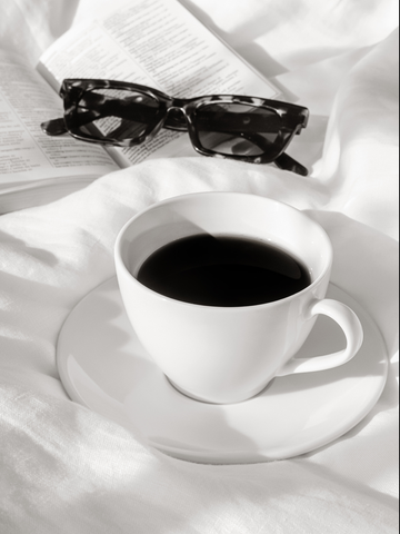 Monochromes Poster mit Kaffeetasse, Buch und Sonnenbrille auf einem Bett.