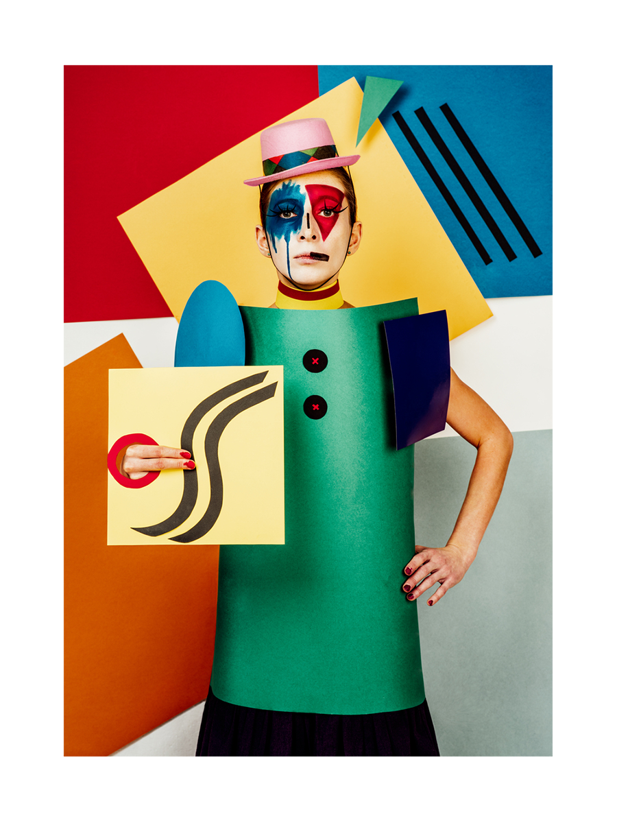 Person in einem farbenfrohen, geometrischen Kostüm mit rot-blau bemaltem Gesicht vor einem Hintergrund aus großen, farbigen Formen.