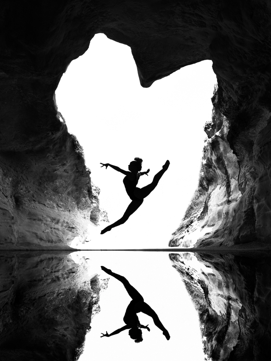 Schwarzweiß-Fotografie einer Tänzerin, die in einer Höhle springt, mit Spiegelung im Wasser.