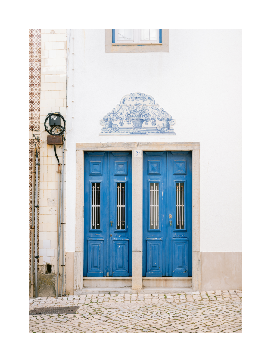 Fotografie von zwei blauen Türen in einem weißen Gebäude mit einem blauen Blumenmuster über der Tür.