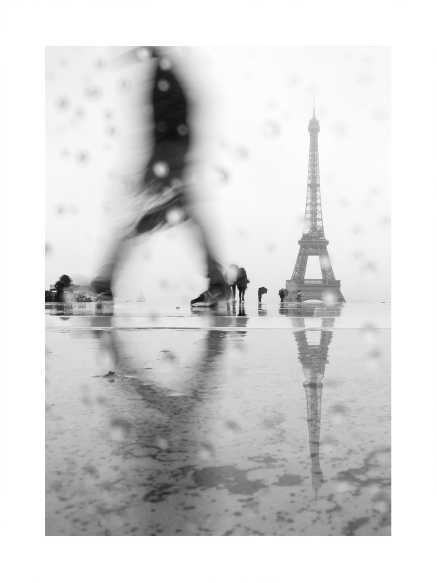 Schwarz-Weiß-Fotografie des Eiffelturms im Regen mit Regentropfen im Vordergrund und einer verschwommenen Person.