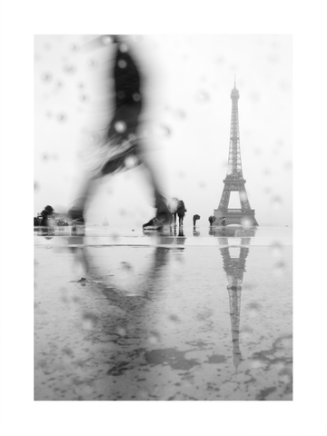 Schwarz-Weiß-Fotografie des Eiffelturms im Regen mit Regentropfen im Vordergrund und einer verschwommenen Person.