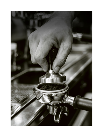 Schwarz-Weiß-Fotografie einer Hand, die Kaffeepulver in einem Siebträger mit einem Tamper presst.