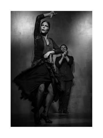Schwarz-Weiß-Foto einer Flamenco-Tänzerin in einem schwarzen Kleid mit einem klatschenden Mann im Hintergrund.