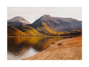 Fotografie von Bergen, die sich in einem See spiegeln, mit einem sandigen Ufer im Vordergrund.