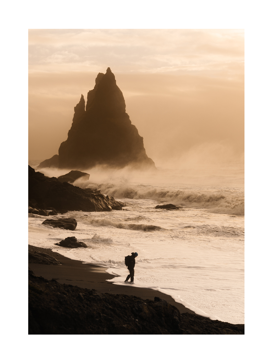 Fotografie eines Reisenden am Strand vor einem Vulkan mit großen Wellen und goldenem Himmel.