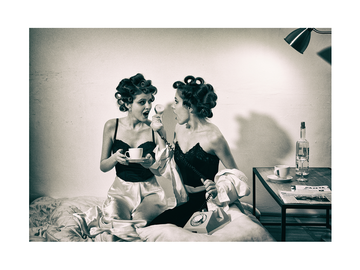 Zwei Frauen mit Lockenwicklern und Kaffeetassen im Bett, eine hält ein Telefon.