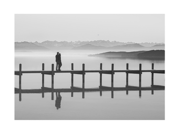 Schwarz-Weiß-Foto eines Paares auf einem Steg über einem stillen See mit Bergen im Hintergrund.